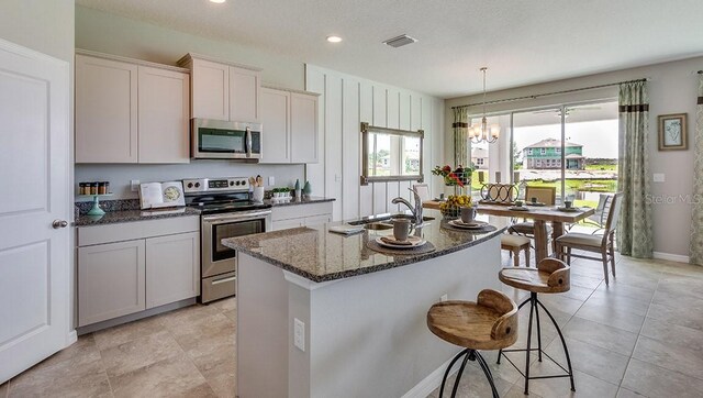 kitchen featuring a kitchen island with sink, dark stone countertops, light tile patterned floors, hanging light fixtures, and appliances with stainless steel finishes