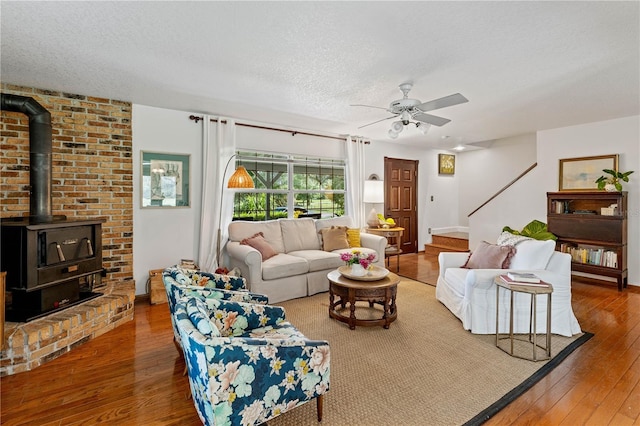 living room featuring ceiling fan, brick wall, a wood stove, hardwood / wood-style flooring, and a textured ceiling