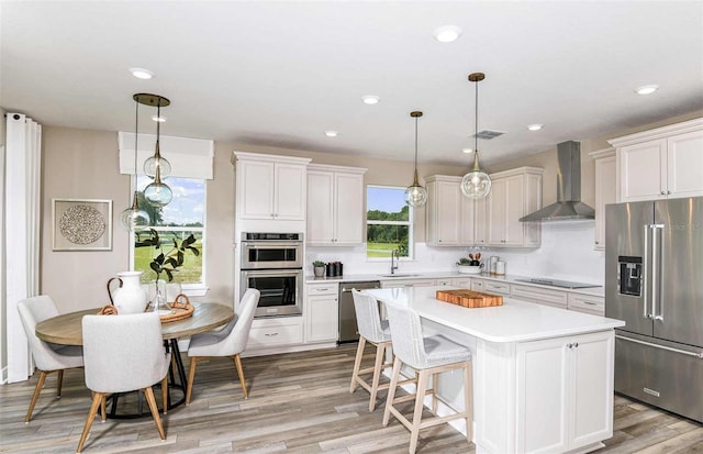 kitchen featuring wall chimney range hood, light wood-type flooring, a kitchen island, hanging light fixtures, and stainless steel appliances