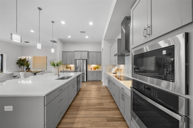 kitchen featuring light wood-style flooring, gray cabinets, stainless steel appliances, wall chimney exhaust hood, and a sink