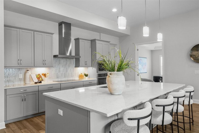 kitchen with an island with sink, oven, gray cabinetry, wall chimney exhaust hood, and tasteful backsplash