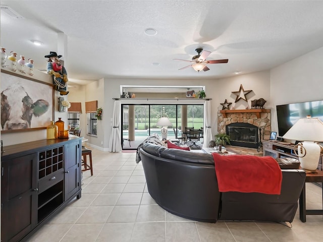 living room with ceiling fan, a stone fireplace, light tile patterned flooring, and a textured ceiling