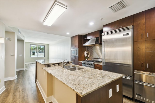 kitchen with built in appliances, light stone countertops, wood-type flooring, backsplash, and a kitchen island with sink