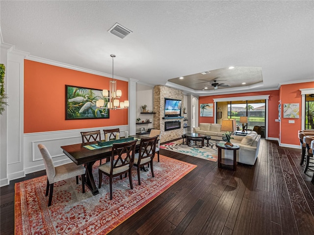 dining space with ceiling fan with notable chandelier, dark hardwood / wood-style flooring, a raised ceiling, a stone fireplace, and a textured ceiling