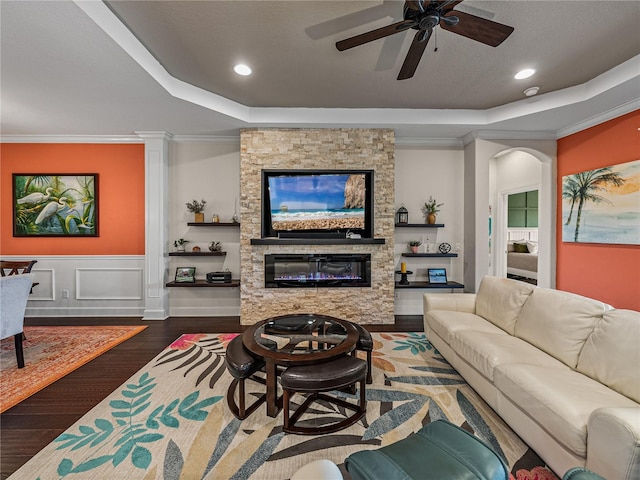 living room with a textured ceiling, ceiling fan, a stone fireplace, a tray ceiling, and dark wood-type flooring