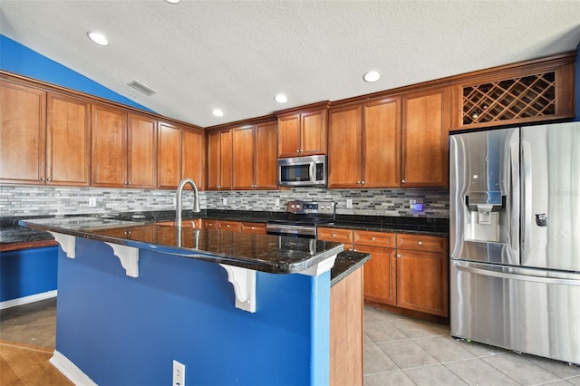 kitchen featuring appliances with stainless steel finishes, decorative backsplash, a kitchen bar, a center island with sink, and dark stone countertops