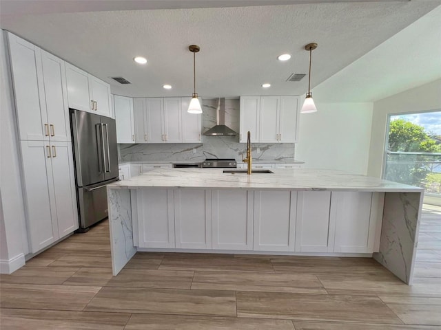 kitchen featuring white cabinetry, appliances with stainless steel finishes, decorative light fixtures, wall chimney range hood, and light stone counters