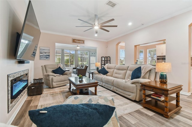 living room featuring visible vents, ornamental molding, light wood-style floors, arched walkways, and a glass covered fireplace