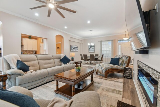 living room featuring ceiling fan with notable chandelier, crown molding, light hardwood / wood-style floors, and a stone fireplace