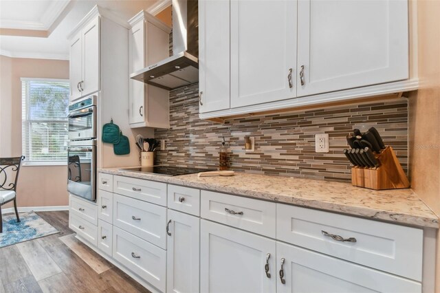 kitchen featuring light hardwood / wood-style flooring, stainless steel double oven, wall chimney exhaust hood, decorative backsplash, and white cabinetry