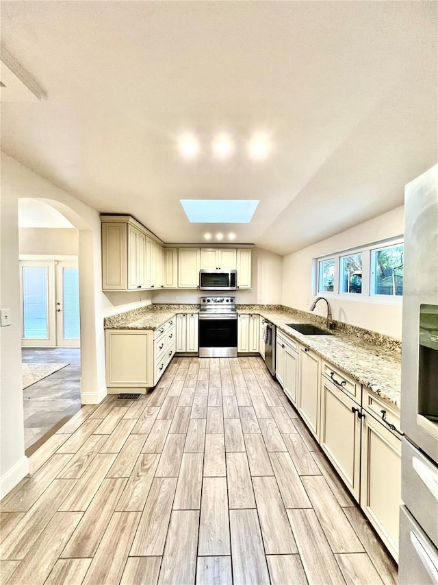 kitchen featuring cream cabinets, sink, vaulted ceiling with skylight, light stone countertops, and appliances with stainless steel finishes