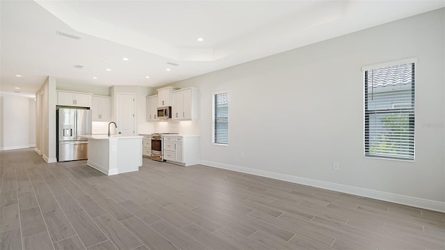 kitchen with appliances with stainless steel finishes, white cabinetry, a raised ceiling, an island with sink, and decorative backsplash