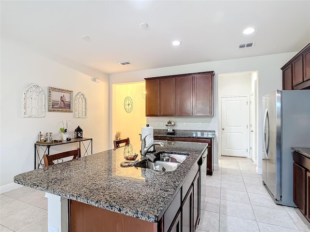 kitchen with dark stone countertops, a center island with sink, stainless steel refrigerator, sink, and light tile patterned floors