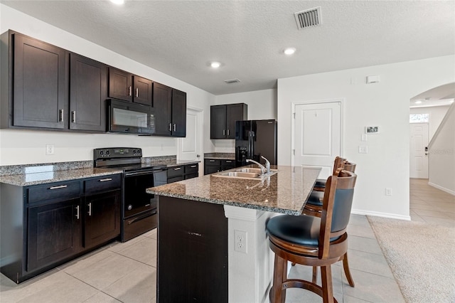 kitchen with an island with sink, black appliances, dark stone counters, light tile patterned flooring, and sink