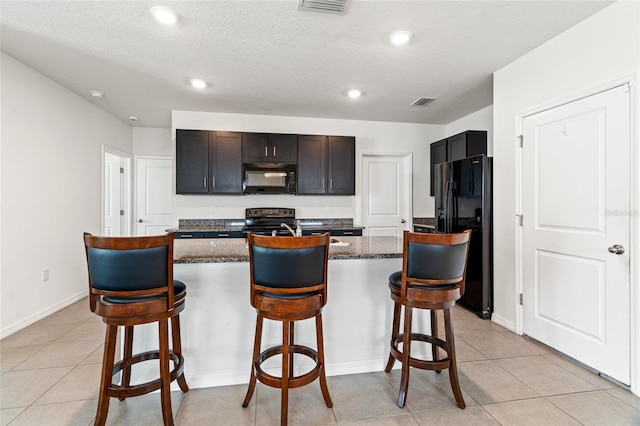 kitchen featuring a breakfast bar area, black appliances, an island with sink, dark stone counters, and a textured ceiling