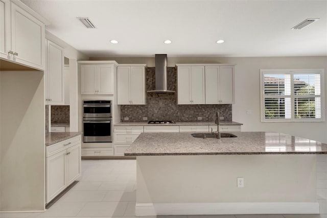 kitchen featuring a kitchen island with sink, white cabinets, wall chimney range hood, sink, and stainless steel double oven