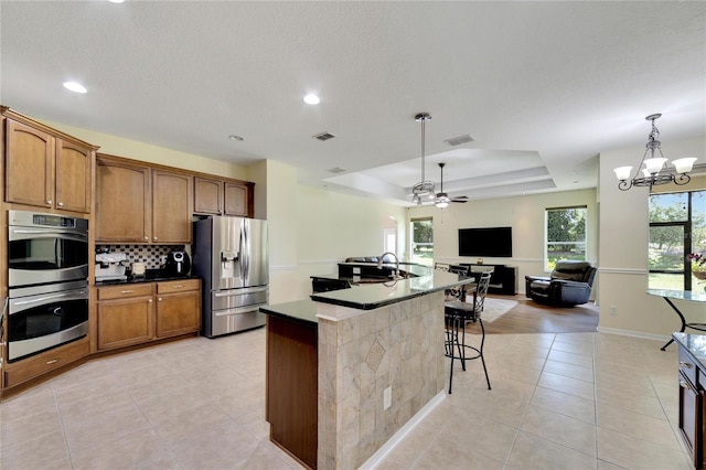 kitchen featuring ceiling fan with notable chandelier, pendant lighting, appliances with stainless steel finishes, a tray ceiling, and an island with sink