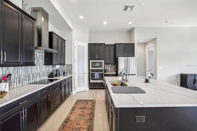 kitchen featuring a center island with sink, appliances with stainless steel finishes, decorative backsplash, light stone counters, and wall chimney range hood