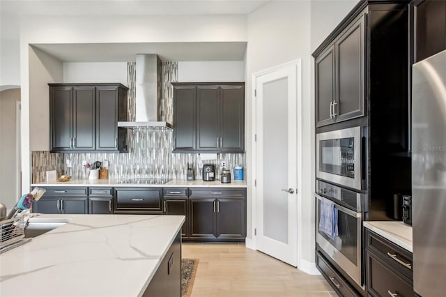 kitchen featuring light stone counters, stainless steel appliances, decorative backsplash, wall chimney exhaust hood, and light wood finished floors