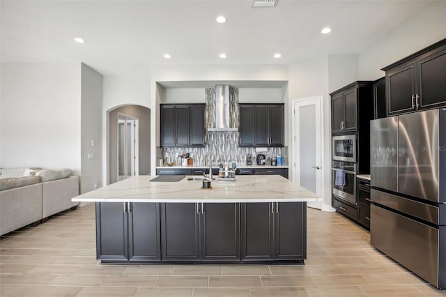 kitchen featuring light stone counters, arched walkways, tasteful backsplash, appliances with stainless steel finishes, and wall chimney range hood