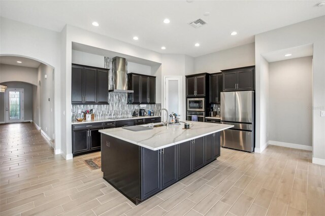 kitchen featuring appliances with stainless steel finishes, light stone counters, wall chimney range hood, an island with sink, and tasteful backsplash