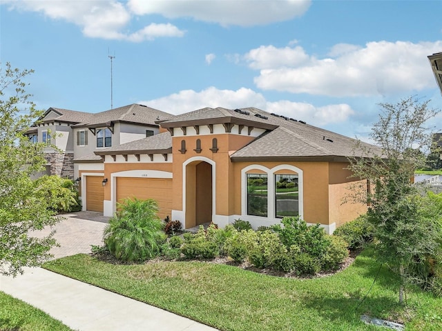 view of front facade featuring decorative driveway, stucco siding, a shingled roof, a front yard, and a garage
