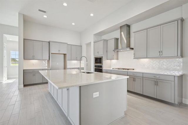 kitchen featuring a center island with sink, sink, wall chimney exhaust hood, gray cabinets, and appliances with stainless steel finishes