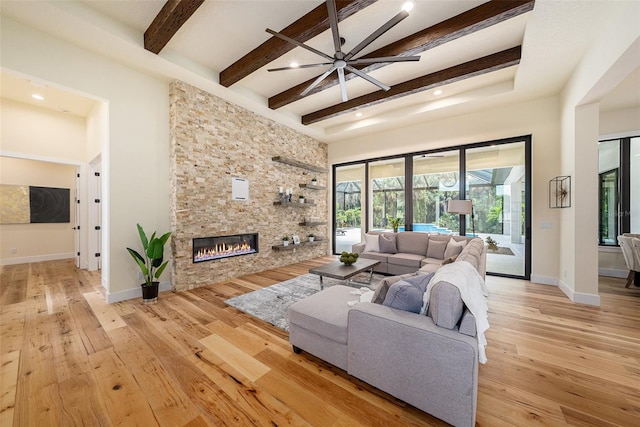 living room with ceiling fan, beam ceiling, a towering ceiling, light hardwood / wood-style floors, and a stone fireplace