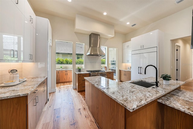 kitchen featuring white cabinetry, sink, exhaust hood, light stone counters, and a center island with sink