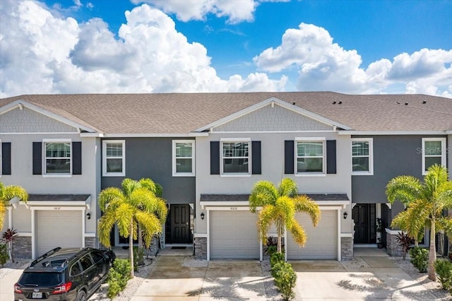 multi unit property with driveway, a shingled roof, stone siding, an attached garage, and stucco siding