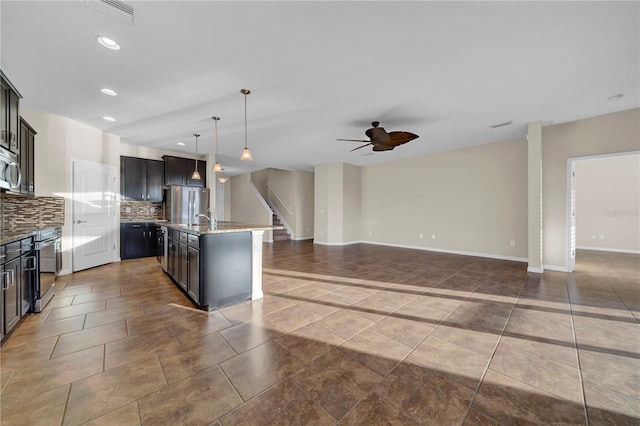 kitchen with pendant lighting, appliances with stainless steel finishes, a kitchen island with sink, light stone countertops, and decorative backsplash