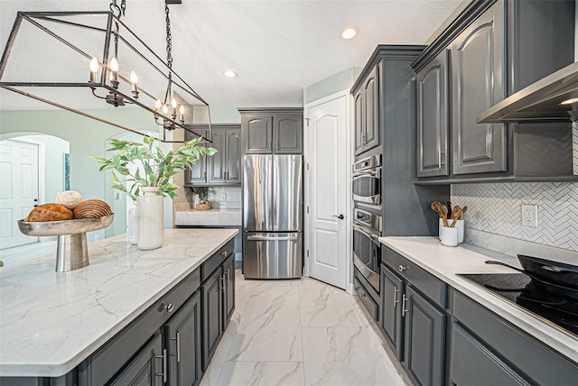 kitchen with a textured ceiling, stainless steel appliances, light stone counters, an inviting chandelier, and wall chimney exhaust hood