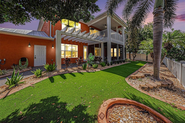 back house at dusk featuring a balcony, a pergola, a lawn, and a patio