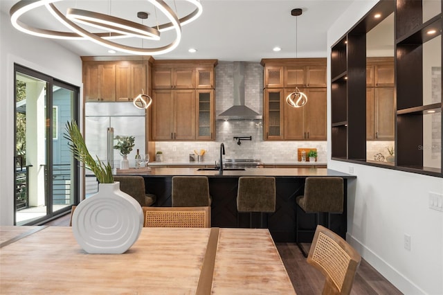 kitchen featuring decorative backsplash, wall chimney exhaust hood, brown cabinets, stainless steel built in fridge, and a sink