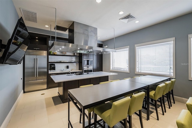 kitchen featuring backsplash, a kitchen island with sink, built in appliances, hanging light fixtures, and range hood