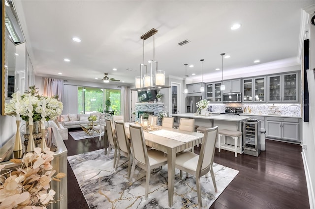 dining area featuring dark wood-type flooring, ceiling fan, wine cooler, and a stone fireplace