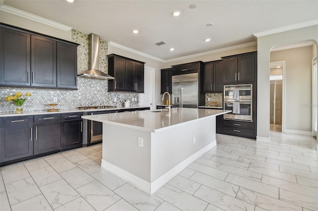 kitchen with crown molding, stainless steel appliances, sink, wall chimney exhaust hood, and a center island with sink