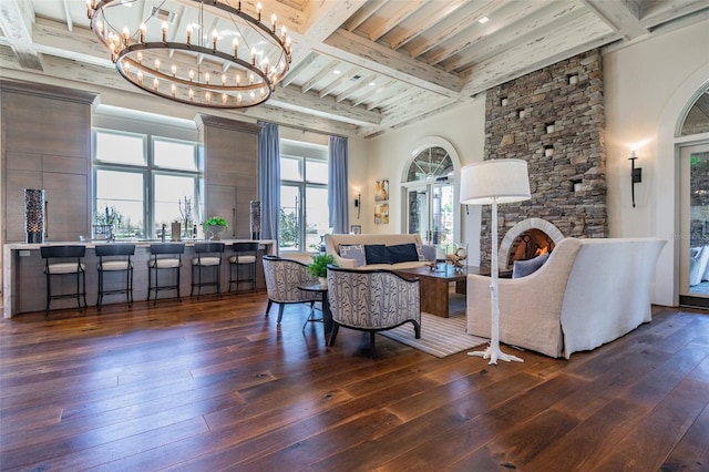 living room with a stone fireplace, dark wood-type flooring, an inviting chandelier, and beamed ceiling