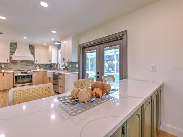 kitchen with french doors, appliances with stainless steel finishes, light wood-type flooring, custom exhaust hood, and decorative backsplash