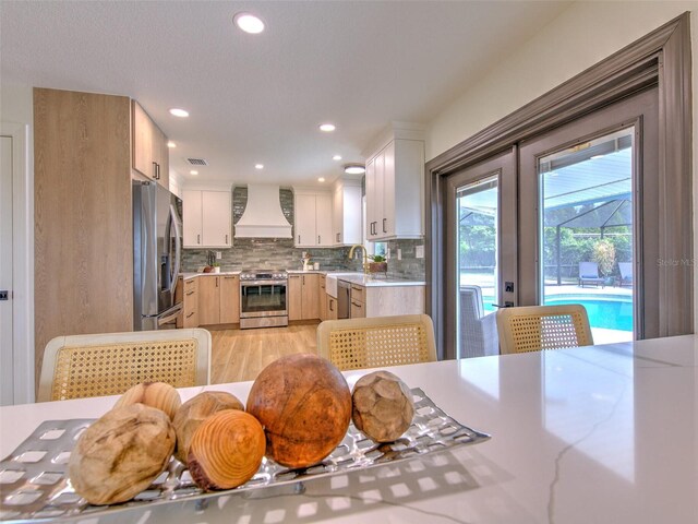 kitchen with tasteful backsplash, light brown cabinetry, light wood-style floors, appliances with stainless steel finishes, and custom exhaust hood