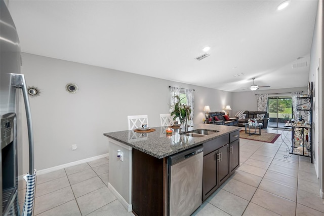 kitchen with a kitchen island with sink, dark stone countertops, dark brown cabinets, stainless steel appliances, and ceiling fan