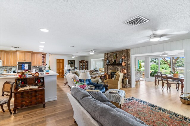 living room featuring ceiling fan, a brick fireplace, a textured ceiling, and light hardwood / wood-style flooring