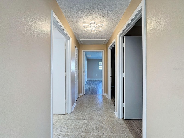 hall featuring attic access, light tile patterned flooring, baseboards, and a textured ceiling