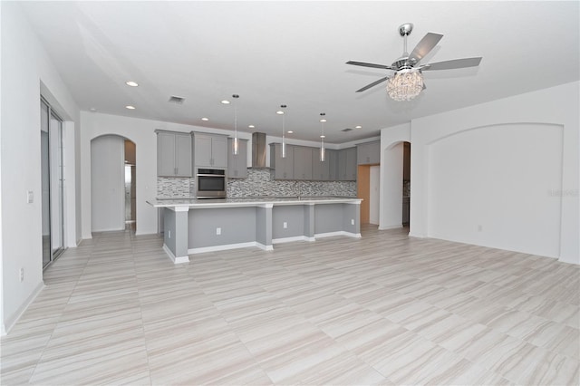 kitchen featuring pendant lighting, a kitchen island with sink, wall chimney range hood, gray cabinets, and decorative backsplash