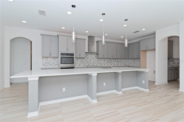 kitchen with wall chimney exhaust hood, gray cabinetry, oven, decorative backsplash, and a spacious island