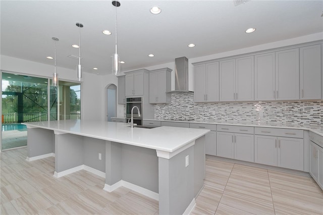 kitchen featuring an island with sink, decorative light fixtures, wall chimney exhaust hood, backsplash, and gray cabinets