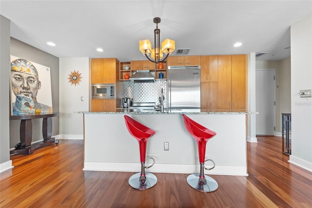 kitchen with built in appliances, dark hardwood / wood-style floors, pendant lighting, stone countertops, and a breakfast bar