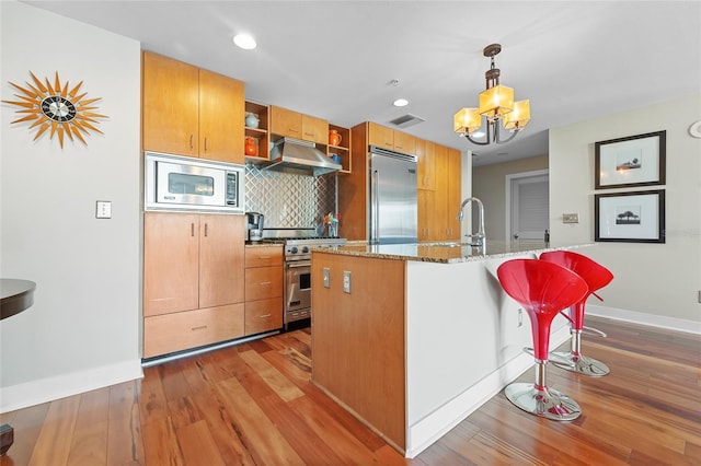 kitchen featuring light wood-type flooring, stone countertops, built in appliances, pendant lighting, and a breakfast bar