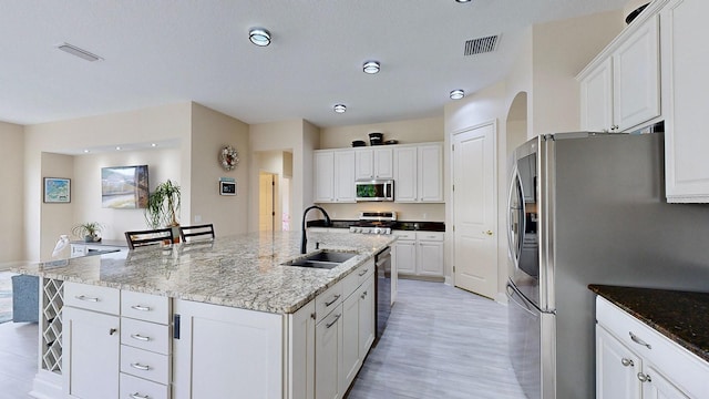 kitchen with sink, appliances with stainless steel finishes, light stone counters, white cabinets, and a center island with sink