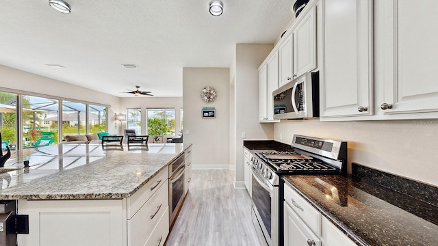 kitchen with appliances with stainless steel finishes, open floor plan, dark stone counters, white cabinetry, and light wood finished floors